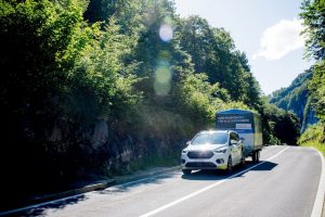 Ein weißer PKW zieht einen großen Anhänger auf einer Landstraße durch einen Wald.