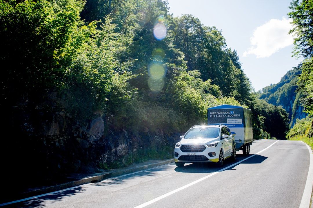 Ein weißer PKW zieht einen großen Anhänger auf einer Landstraße durch einen Wald.